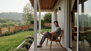 Een man van midden dertig zit ontspannen op een houten veranda bij een tuin in het vroege ochtendlicht