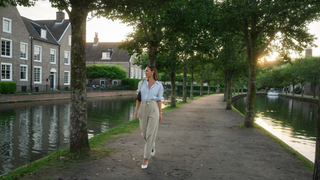 Een vrouw van begin dertig loopt ontspannen over een rustig pad langs een gracht met bomen in het zachte vroege avondlicht