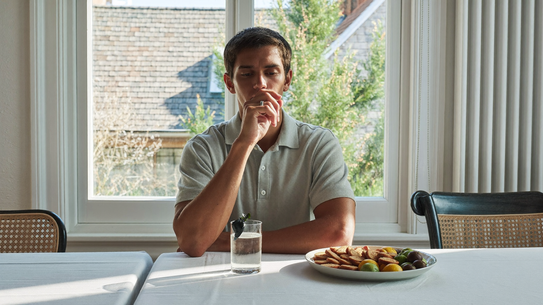 Een jonge man zit op een zondagochtend aan een tafel bij het raam met een glas water en kijkt rustig naar buiten
