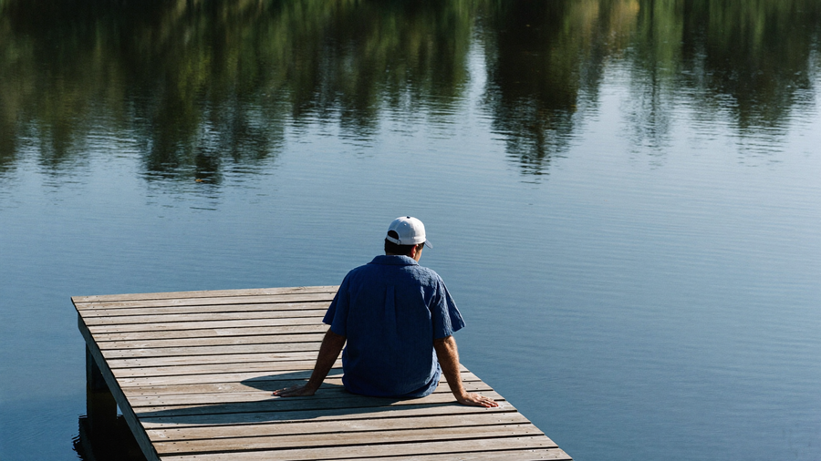Een man van rond de dertig zit in de ochtend op een steiger aan een meer en kijkt rustig over het water