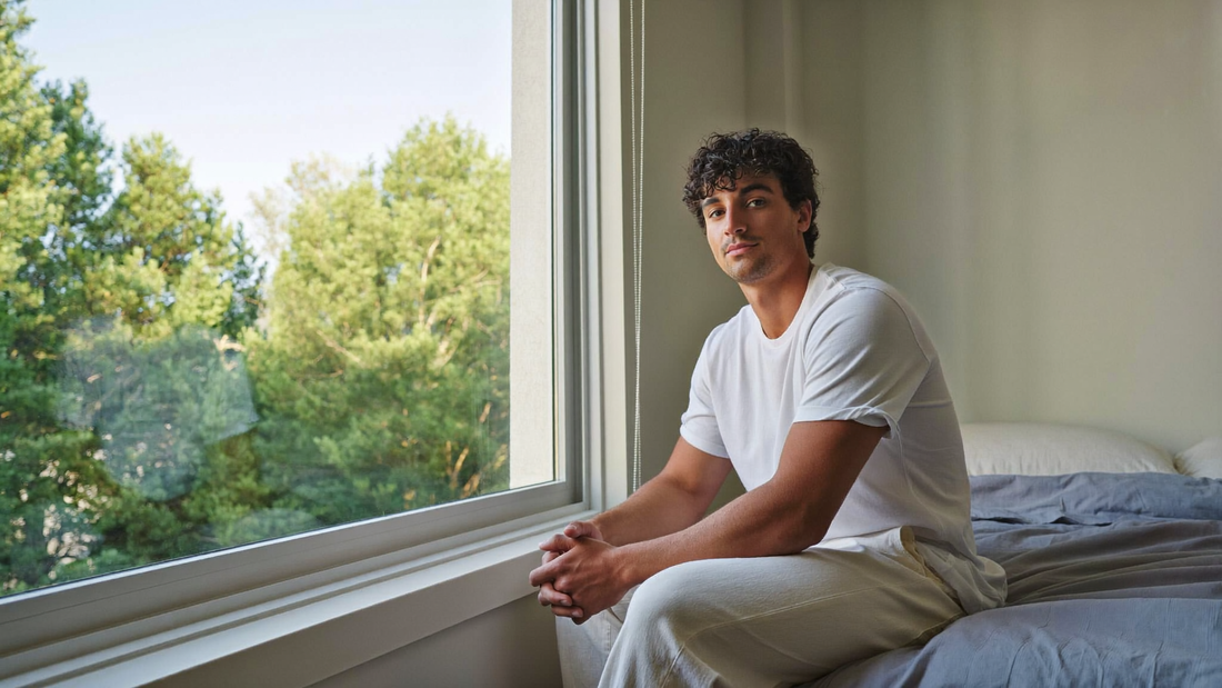 Een man van in de twintig zit ontspannen op de rand van zijn bed bij een open raam in de vroege ochtend, met groene boomtoppen buiten
