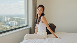 Een vrouw van midden dertig zit ontspannen op de rand van haar bed bij een groot raam op een rustige ochtend, met een ingepakte reistas tegen de muur
