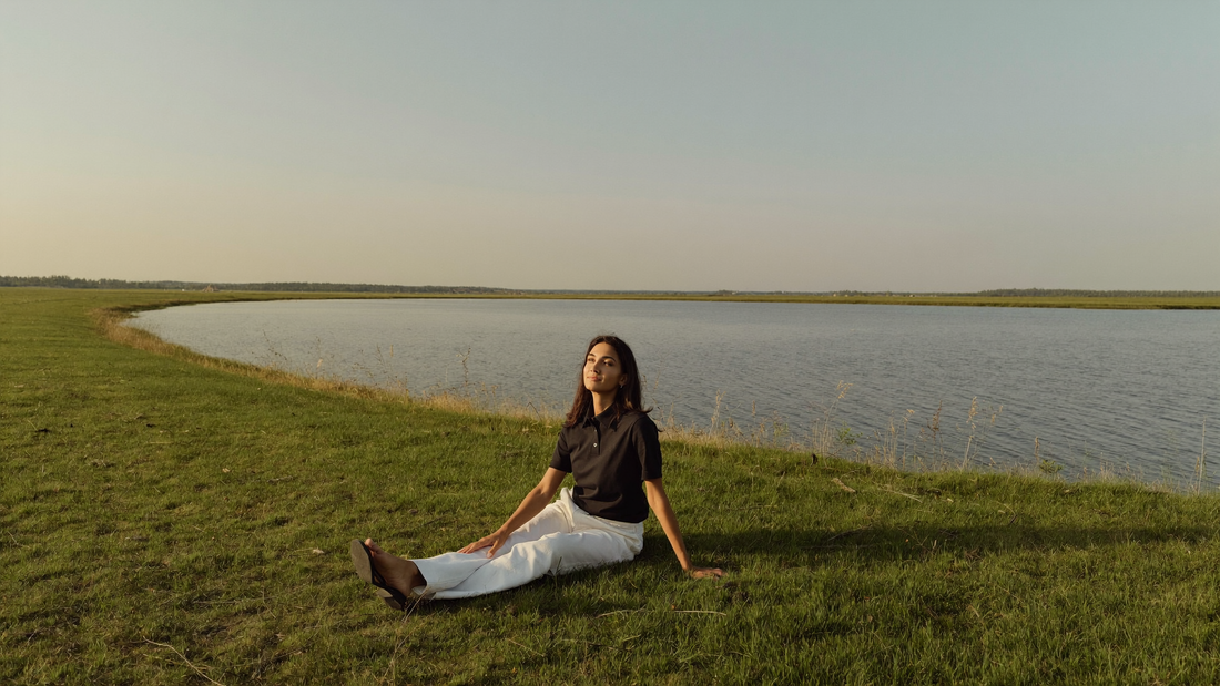 Een vrouw van midden dertig zit ontspannen in het gras op een groene dijk in de vroege avond en kijkt uit over breed, kalm water