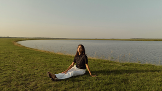 Een vrouw van midden dertig zit ontspannen in het gras op een groene dijk in de vroege avond en kijkt uit over breed, kalm water