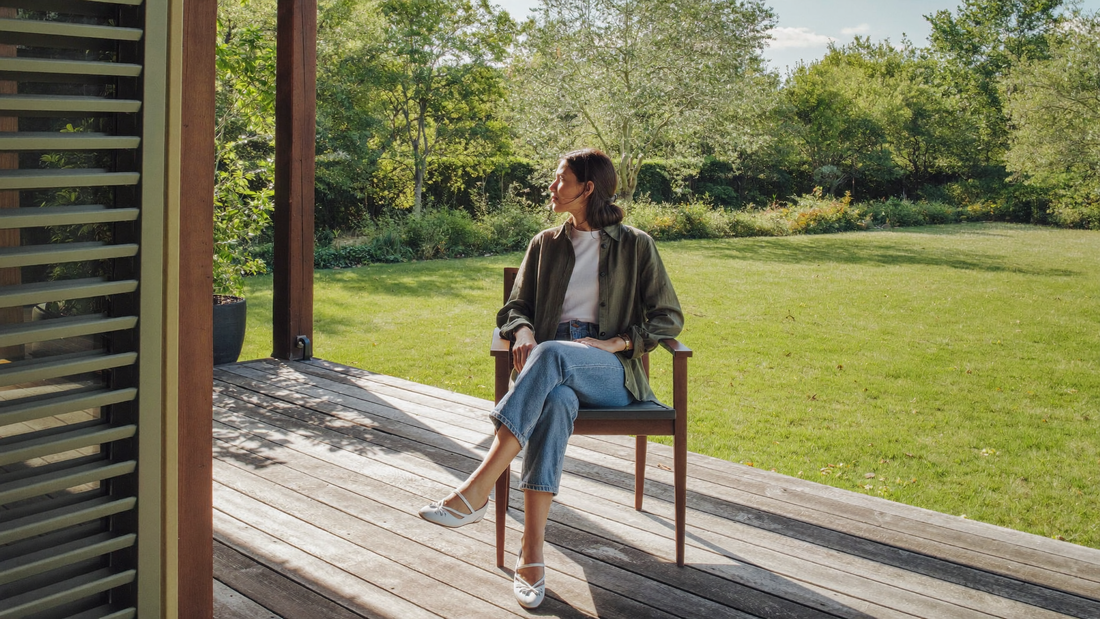 Een vrouw van begin dertig zit ontspannen op een houten veranda en kijkt in de ochtend uit over een groene tuin
