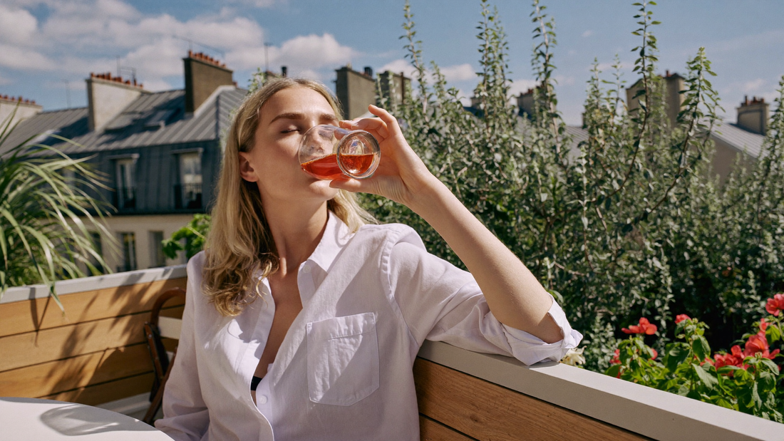 Een jonge vrouw drinkt 's ochtends een glas water op een zonnig balkon met planten en daken op de achtergrond