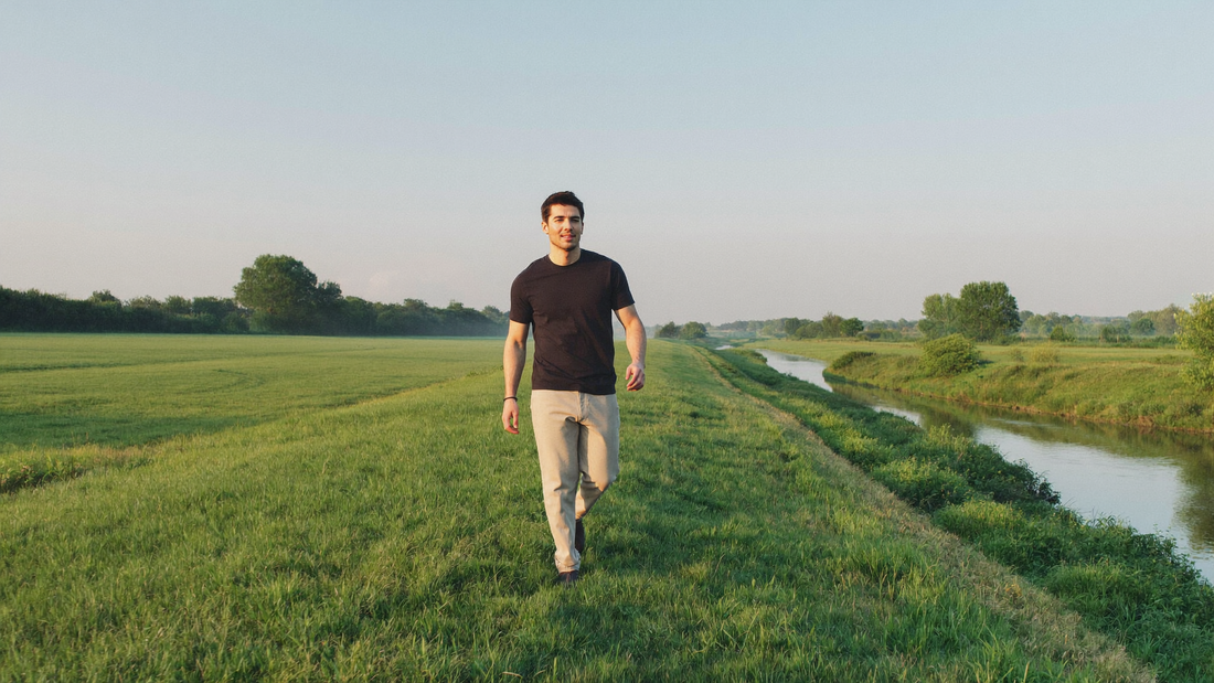 Een man van midden dertig wandelt ontspannen over een grasdijk op een heldere zondagochtend, naast groene weilanden en een rustige rivier