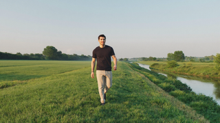 Een man van midden dertig wandelt ontspannen over een grasdijk op een heldere zondagochtend, naast groene weilanden en een rustige rivier