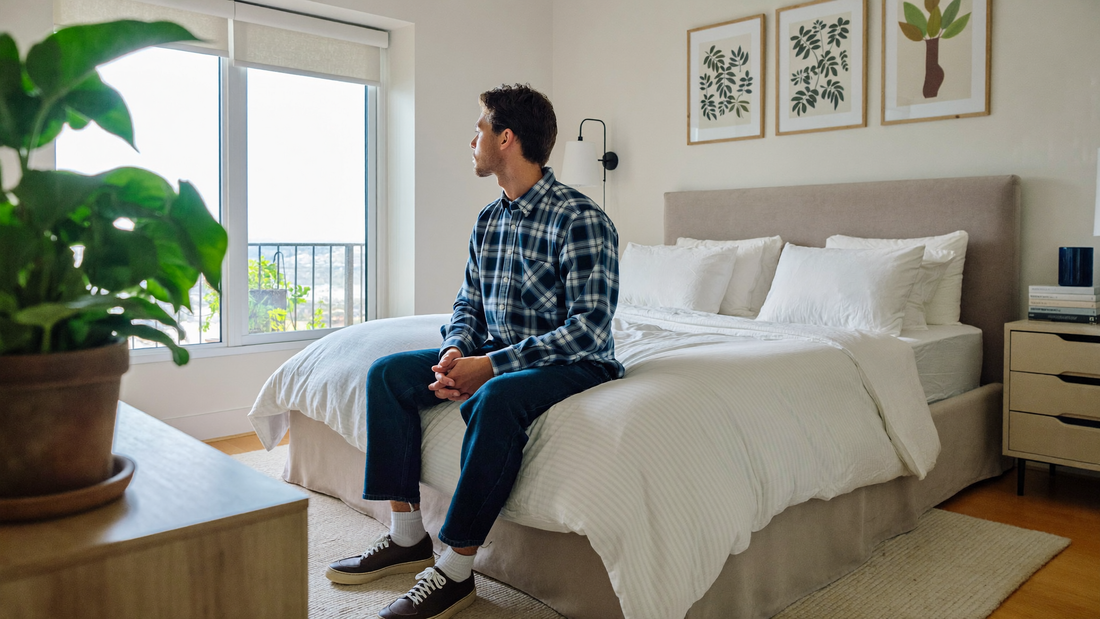 Een man van in de dertig zit 's ochtends op de rand van een opgemaakt bed in een lichte slaapkamer en kijkt rustig uit het raam