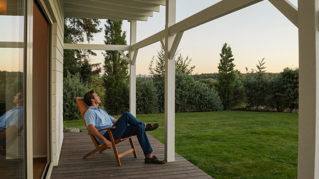 Een man van midden dertig zit ontspannen op een houten veranda in de vroege avond en kijkt uit over een groene tuin