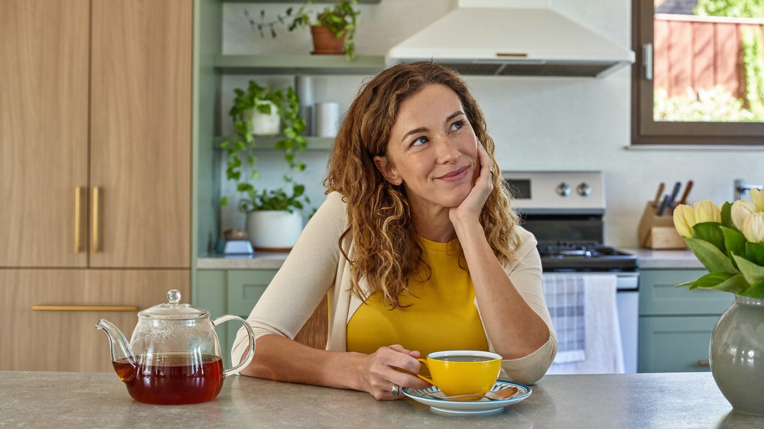 Een vrouw van in de dertig zit aan een keukentafel met een kop thee en kijkt rustig en nadenkend voor zich uit