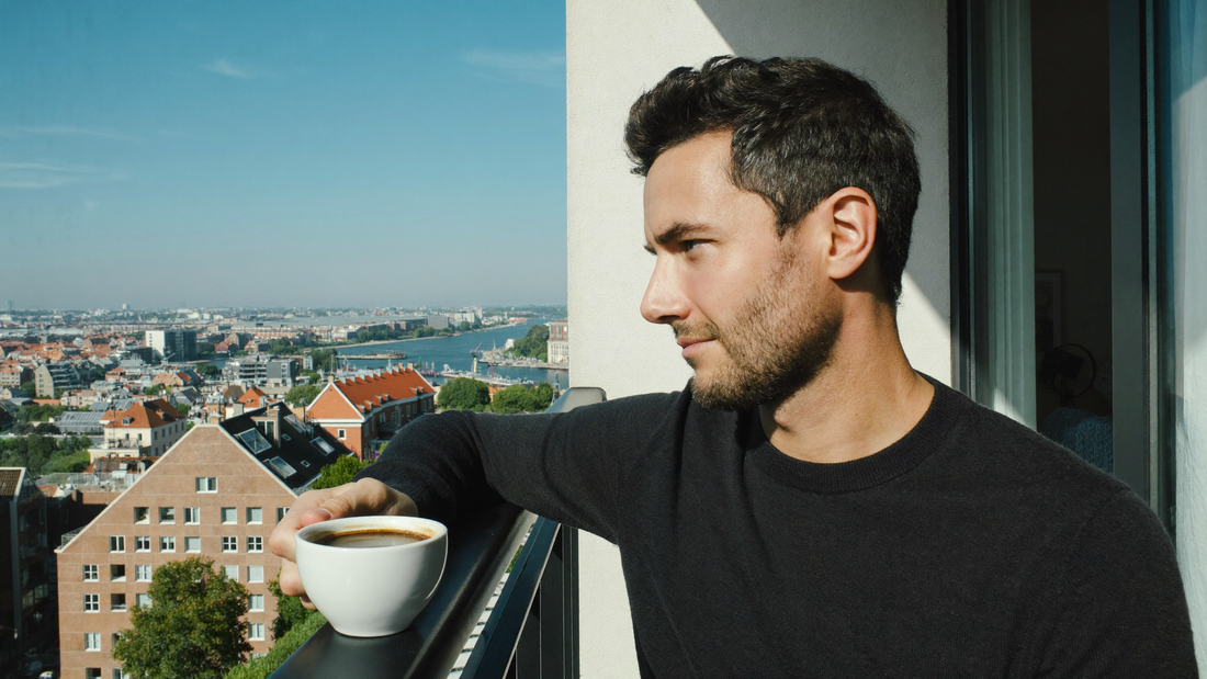 Een man van in de dertig zit op een zonnige ochtend op een balkon met een kop koffie en kijkt rustig over de stad