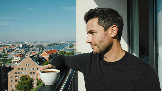 Een man van in de dertig zit op een zonnige ochtend op een balkon met een kop koffie en kijkt rustig over de stad