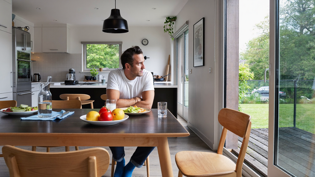 Een man van rond de veertig zit in de ochtend aan een keukentafel bij het raam met een glas water en kijkt rustig naar buiten