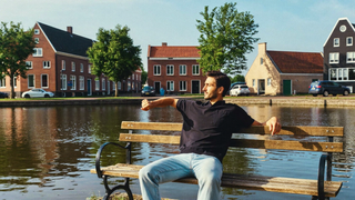Een man van midden dertig zit ontspannen op een houten bank langs een rustige gracht in de vroege ochtend, met bakstenen huizen en groene bomen langs de kade