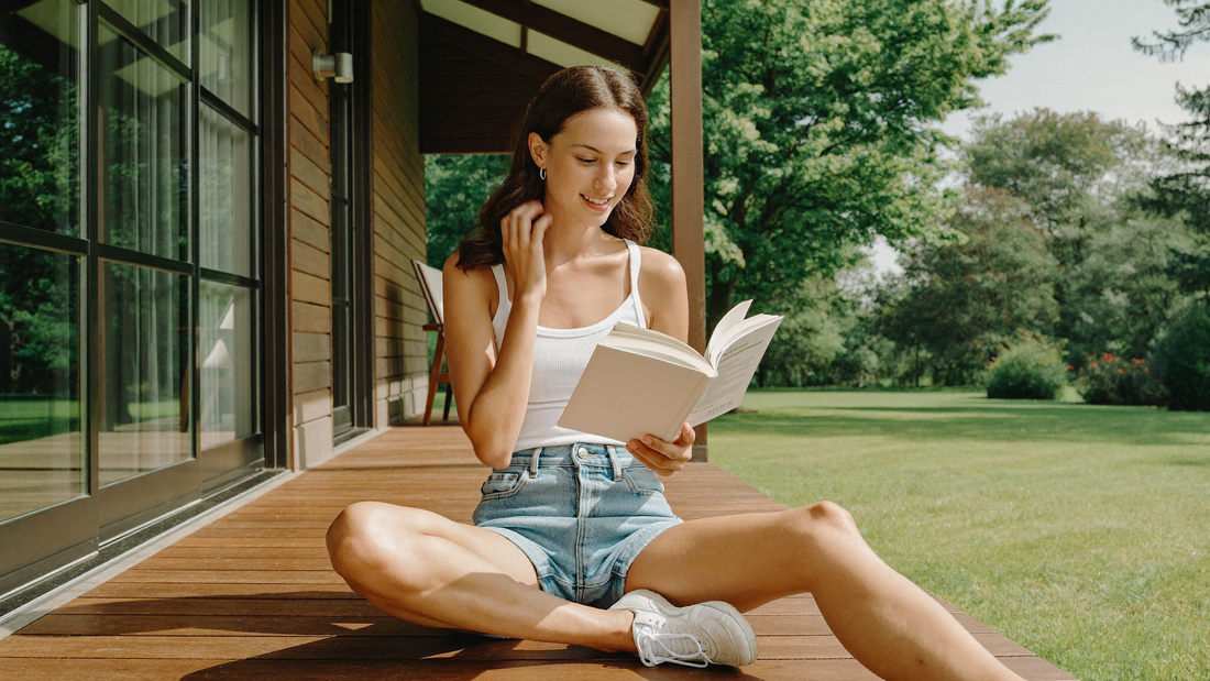 Een jonge vrouw zit in kleermakerszit op een zonnige houten veranda en leest een boek, met een groene tuin op de achtergrond