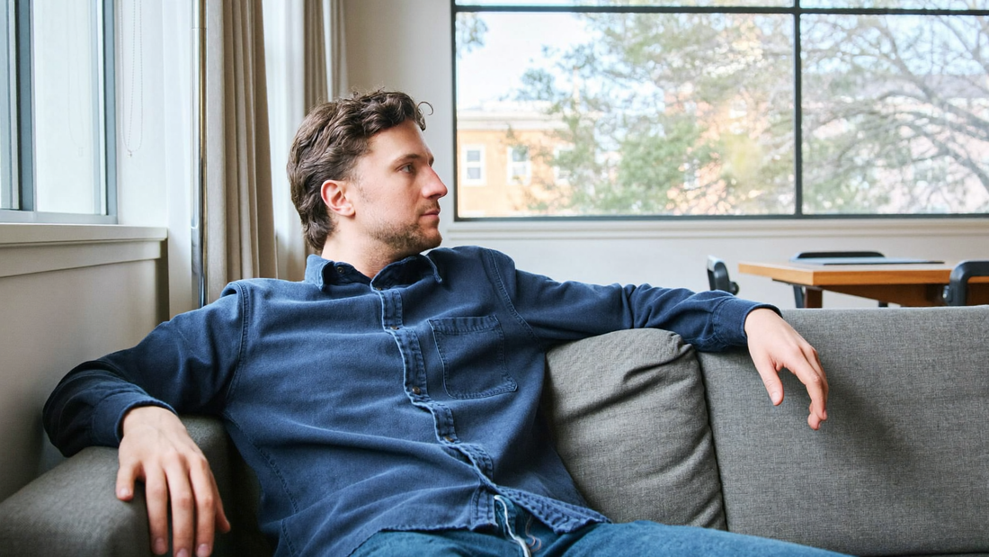 Een man van rond de vijfendertig zit ontspannen op een bank in een lichte woonkamer en kijkt rustig naar buiten