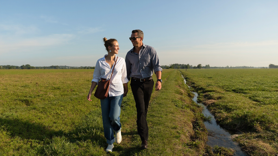 Een vrouw van midden dertig en een man van begin dertig wandelen ontspannen naast elkaar over een grazig dijkpad langs groene weilanden in de ochtend