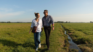 Een vrouw van midden dertig en een man van begin dertig wandelen ontspannen naast elkaar over een grazig dijkpad langs groene weilanden in de ochtend