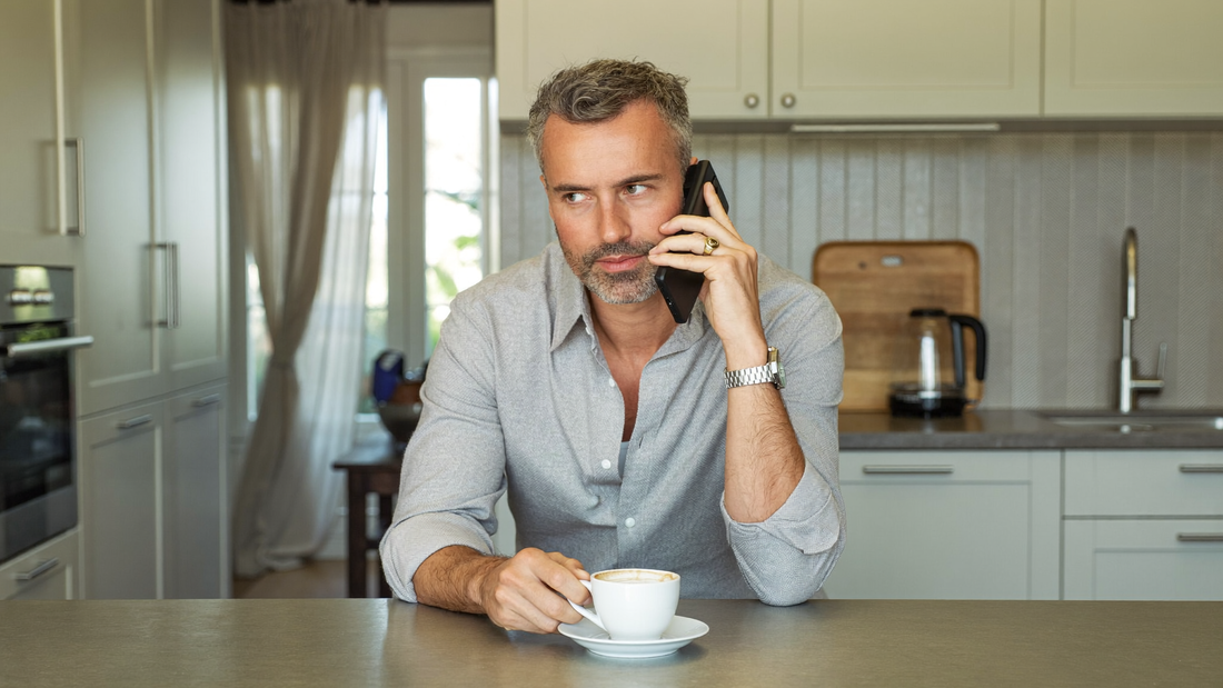 Een man van in de vijftig belt aan de keukentafel met zijn telefoon, een kop koffie naast zich