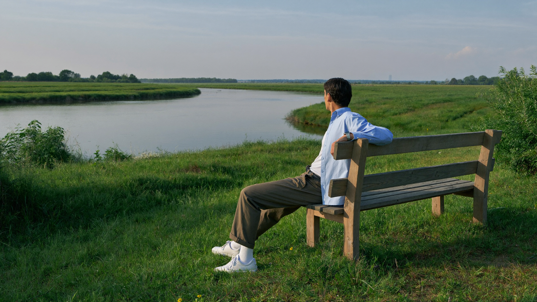 Een man van begin dertig zit ontspannen op een houten bank op een grasdijk en kijkt uit over een brede, kalme rivier in de vroege ochtend