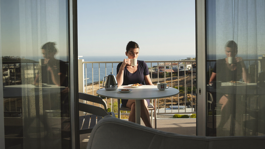 Een vrouw van in de dertig drinkt in de ochtend thee op het balkon van een vakantiehuisje met uitzicht op zee