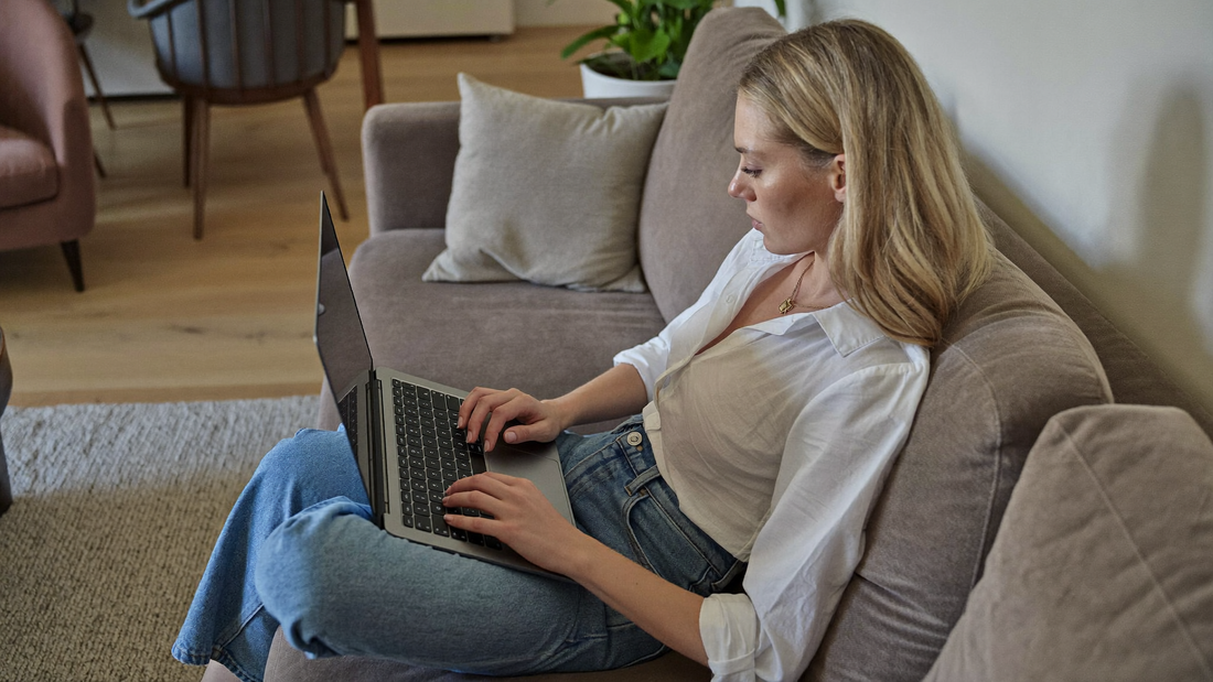 Een vrouw van in de dertig zit op de bank met een laptop op schoot en kijkt aandachtig naar het scherm