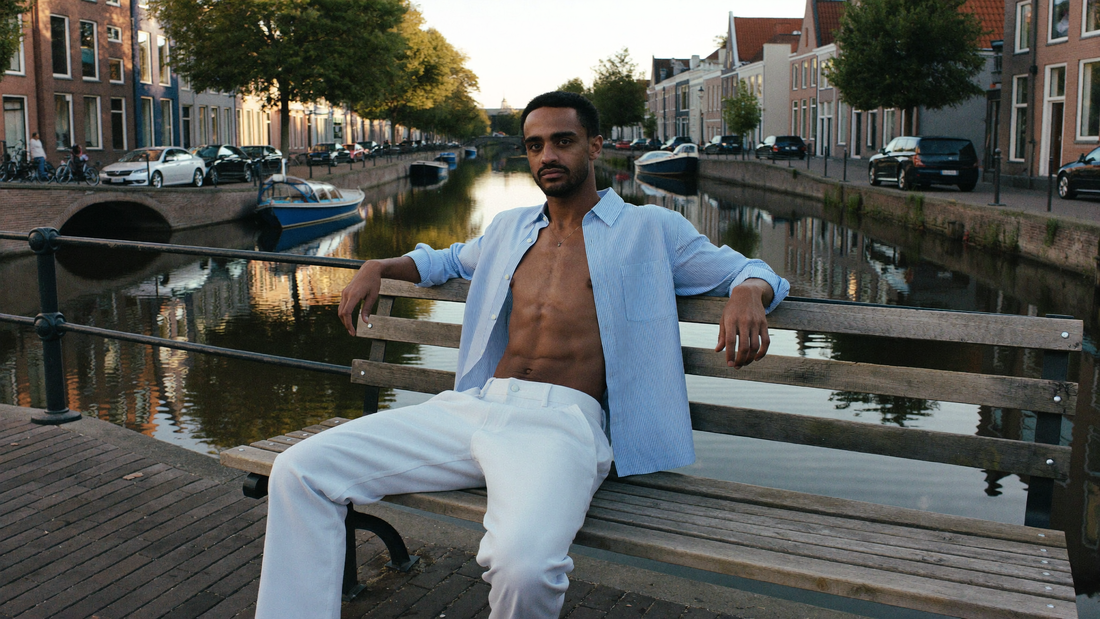 Een man van begin dertig zit ontspannen op een houten bank langs een rustige gracht in de late ochtend