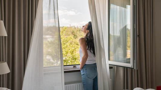 Een vrouw staat uitgerust bij een open slaapkamerraam in het vroege ochtendlicht