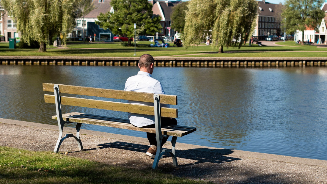 Een man van rond de veertig zit alleen op een bankje aan het water in de ochtend en kijkt rustig voor zich uit