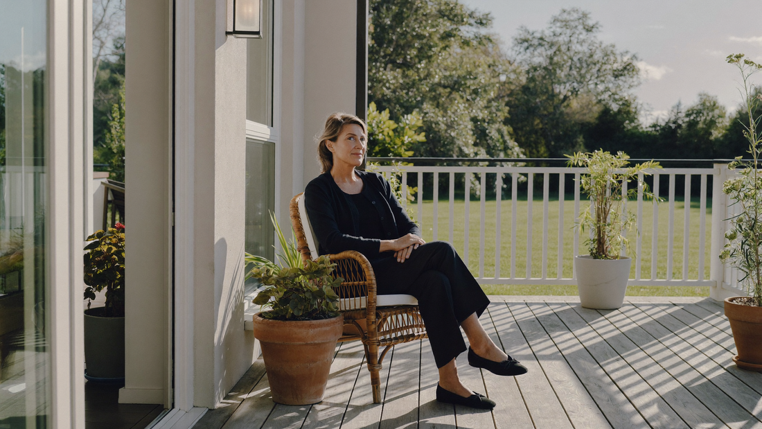 Een vrouw van rond de vijftig zit in een rieten stoel op de veranda van haar huis in het ochtendlicht