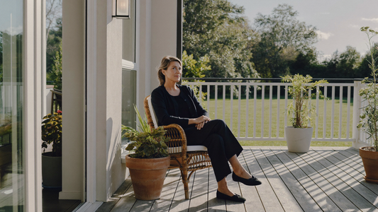 Een vrouw van rond de vijftig zit in een rieten stoel op de veranda van haar huis in het ochtendlicht