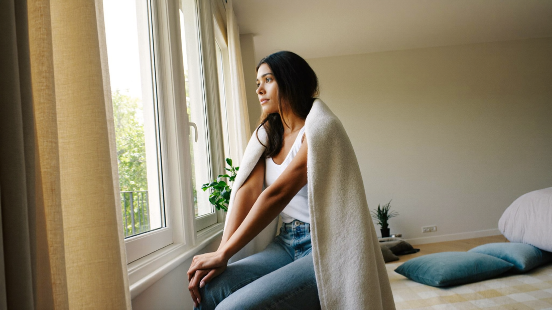 Een vrouw van eind twintig zit in de late ochtend met een deken om bij het raam