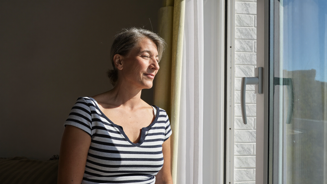 Een vrouw van in de veertig staat bij een open raam in een lichte kamer en laat de ochtendzon op haar gezicht vallen, met een rustige blik