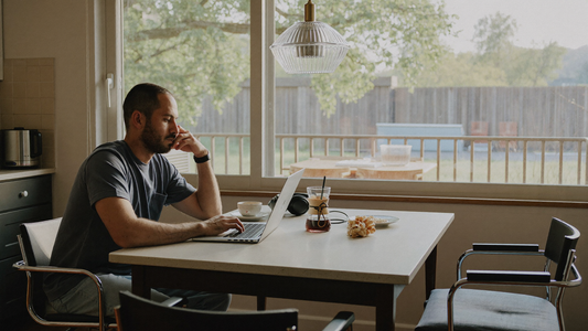 Een man van in de veertig zit thuis aan de keukentafel met een laptop en koptelefoon, rustig en geconcentreerd in het ochtendlicht