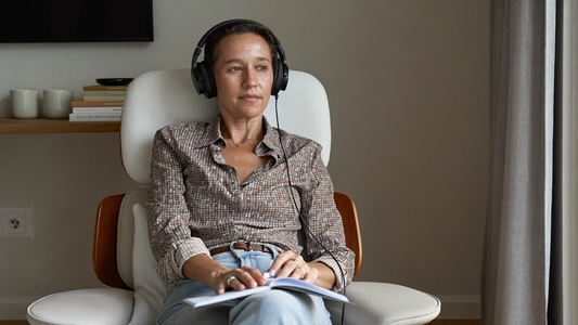 Een vrouw van in de vijftig zit thuis in een comfortabele stoel bij het raam met een koptelefoon op en een notitieboek op schoot