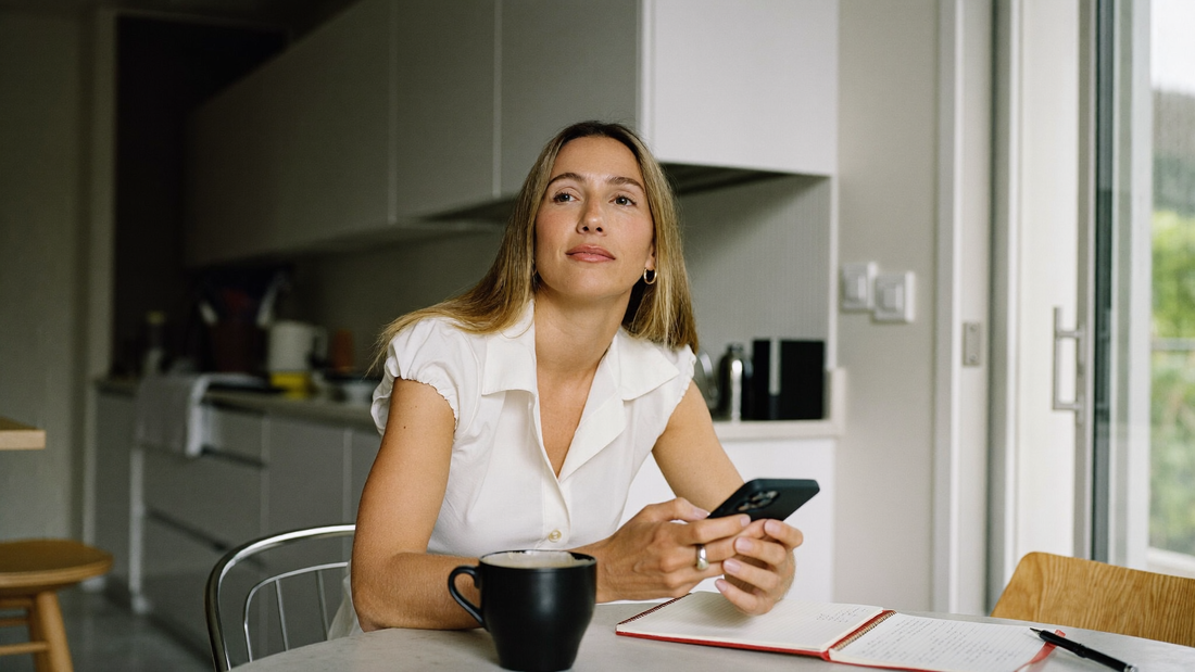 Een vrouw van in de veertig zit aan een keukentafel met haar telefoon en een notitieboekje en kijkt rustig voor zich uit