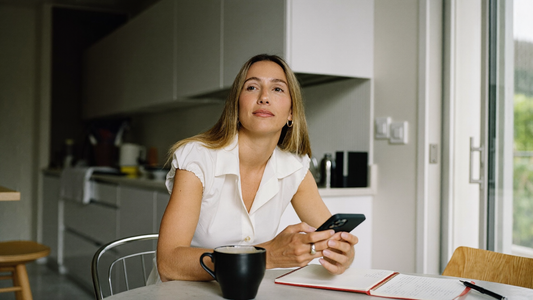 Een vrouw van in de veertig zit aan een keukentafel met haar telefoon en een notitieboekje en kijkt rustig voor zich uit