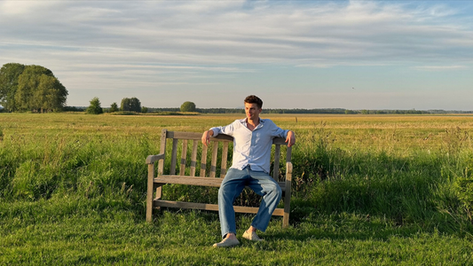 Een man zit ontspannen op een bankje op een dijk en kijkt uit over de polder
