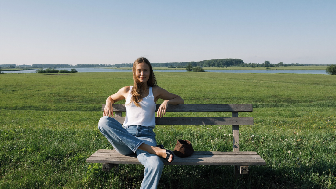 Een vrouw van eind twintig zit ontspannen op een houten bank op een grasdijk op een zonnige doordeweekse ochtend, met weilanden en een rustige rivier achter haar